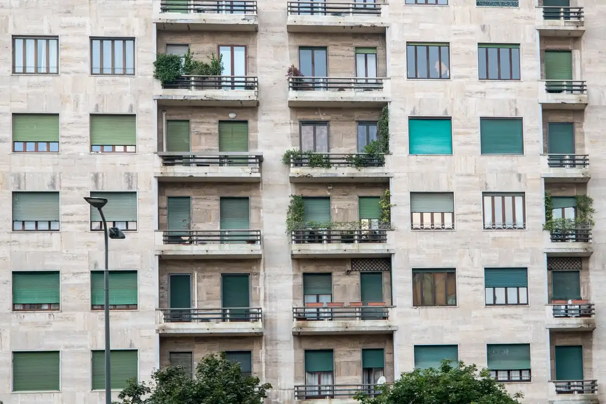 Fachada de un edificio de pisos con balcones de barandilla oscura, persianas verdes y plantas en algunas terrazas